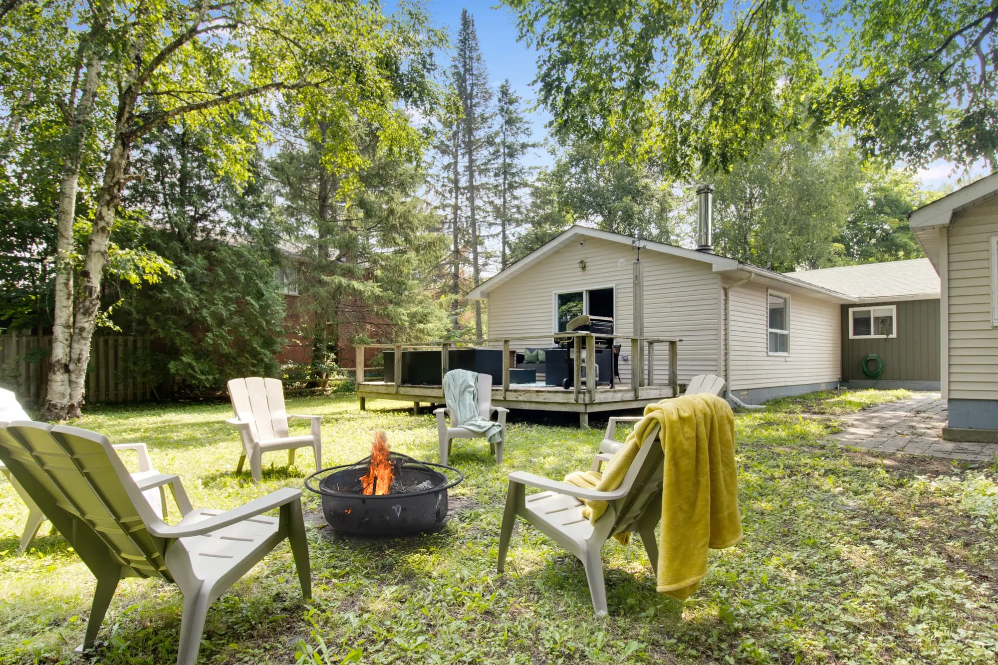 Adirondack chairs and a fire pit on the lawn behind the house, under tall trees